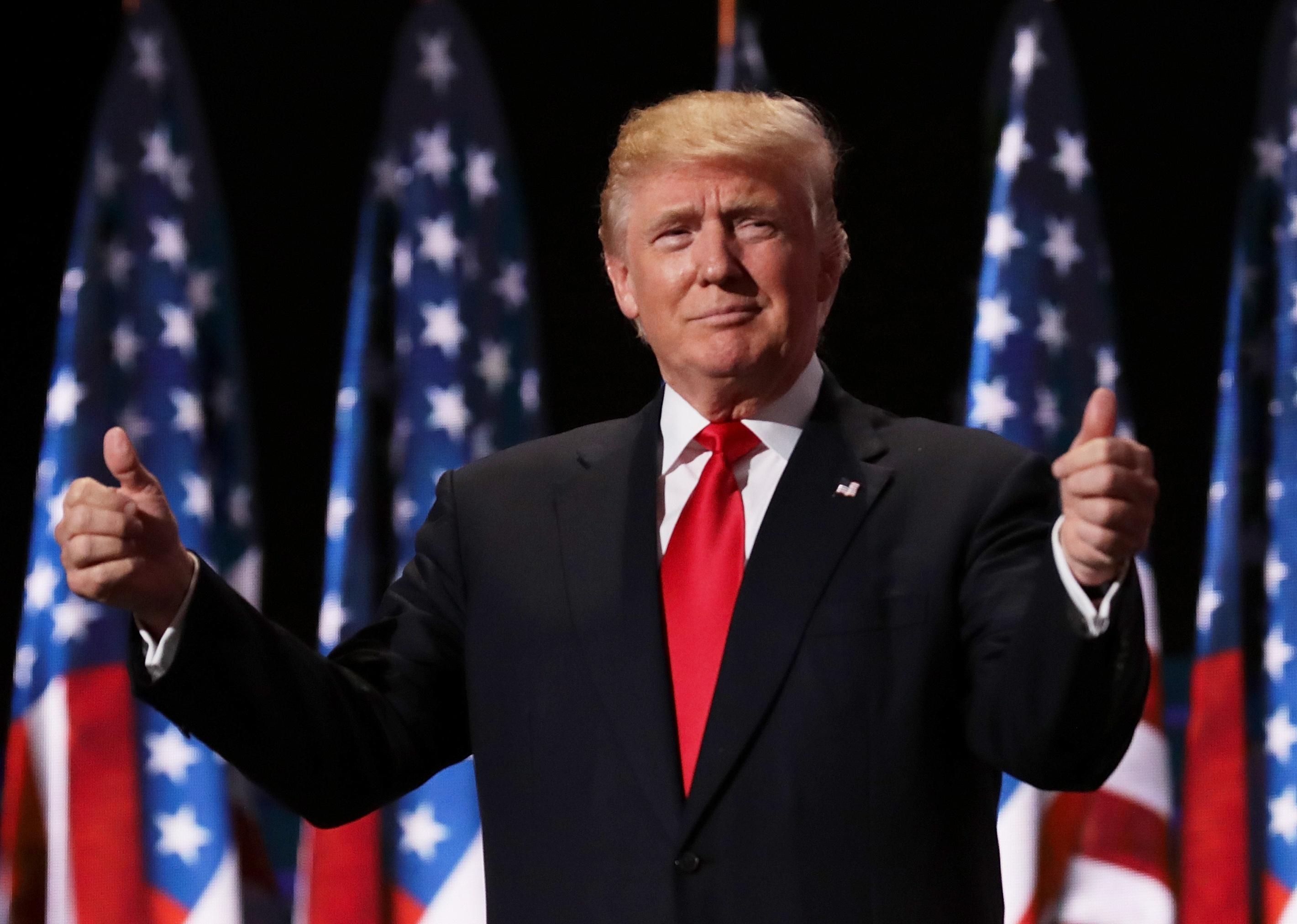 Donald Trump giving two thumbs up onstage in front of American flags.