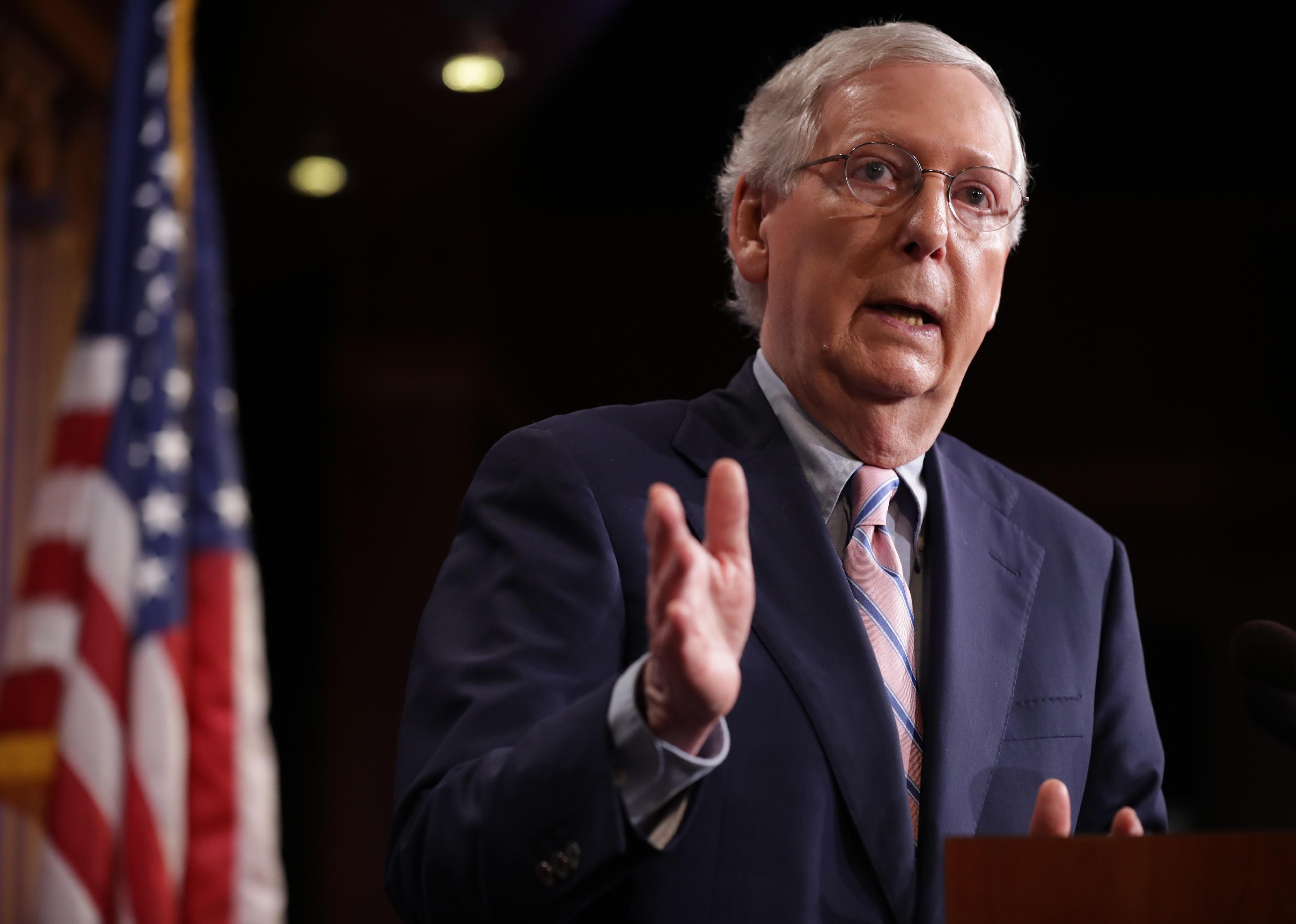 Mitch McConnell, in a suit, speaking at a podium in front of an American flag.