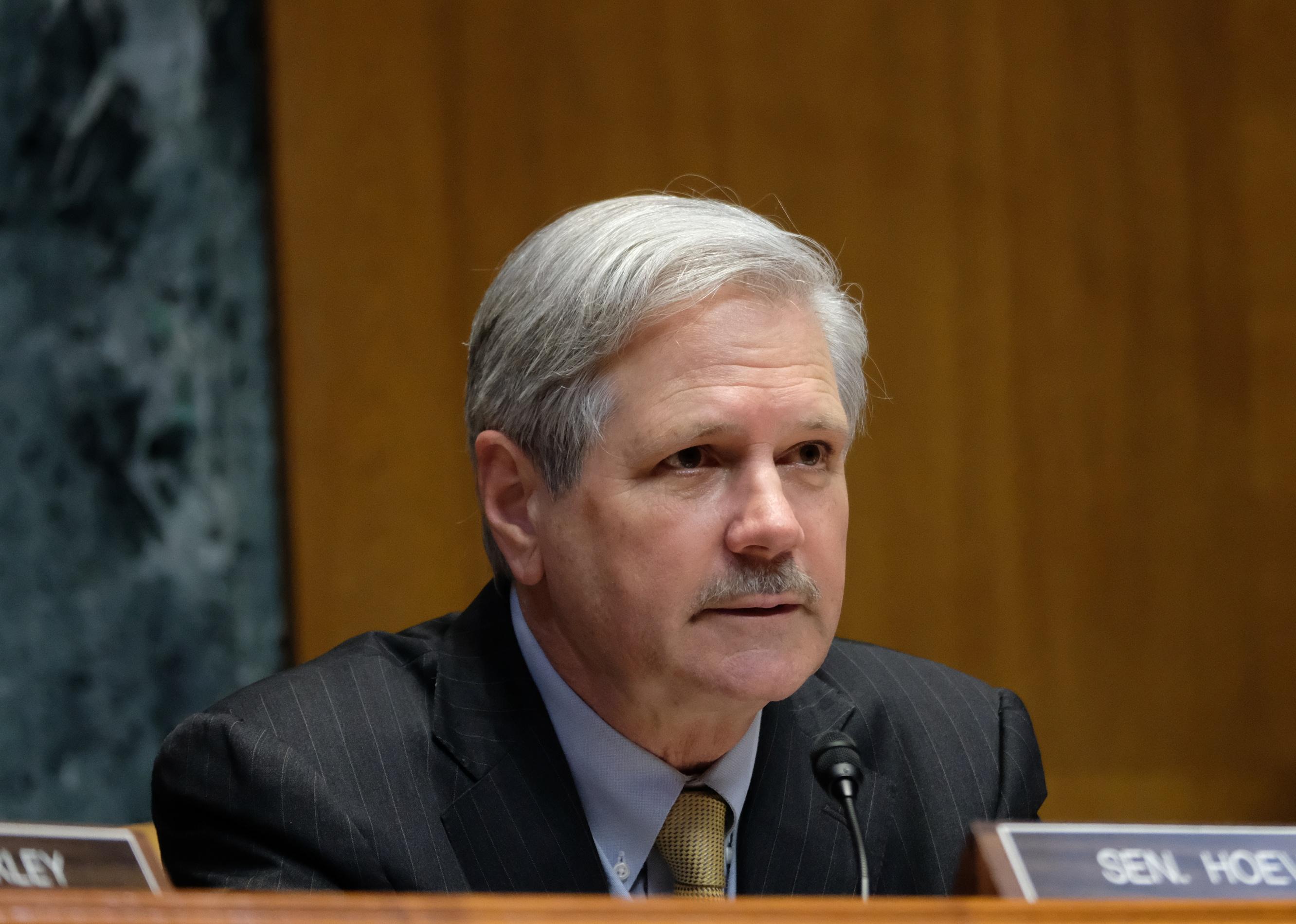 John Hoeven seated in front of his name plate in a suit.