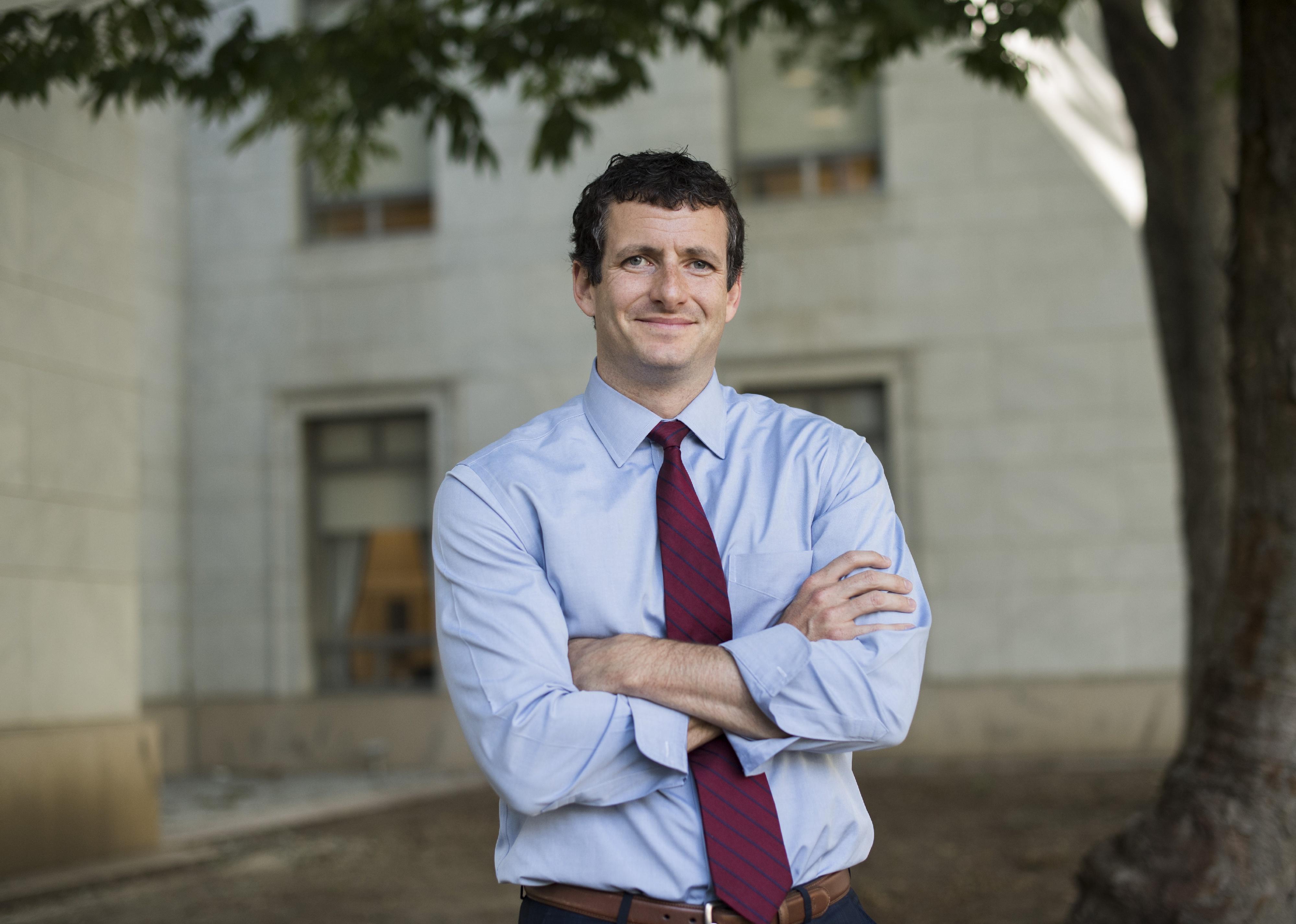 Trey Hollingsworth, in a blue button-down shirt and tie, smiling with his arms crossed in a courtyard.
