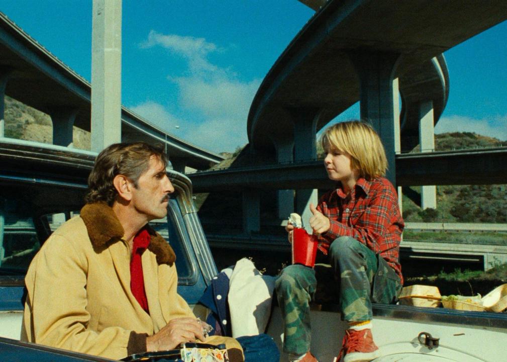 A man and a little boy eat fast food in the back of a pickup truck under an overpass.