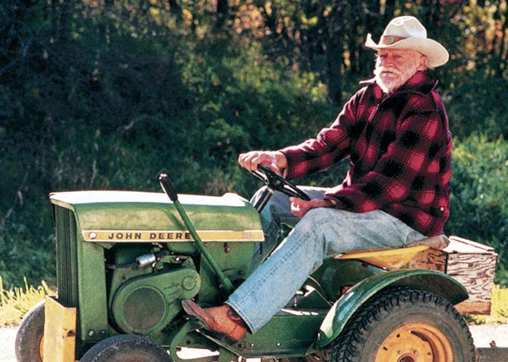 An old man in a cowboy hat riding a John Deere lawnmower on the highway.