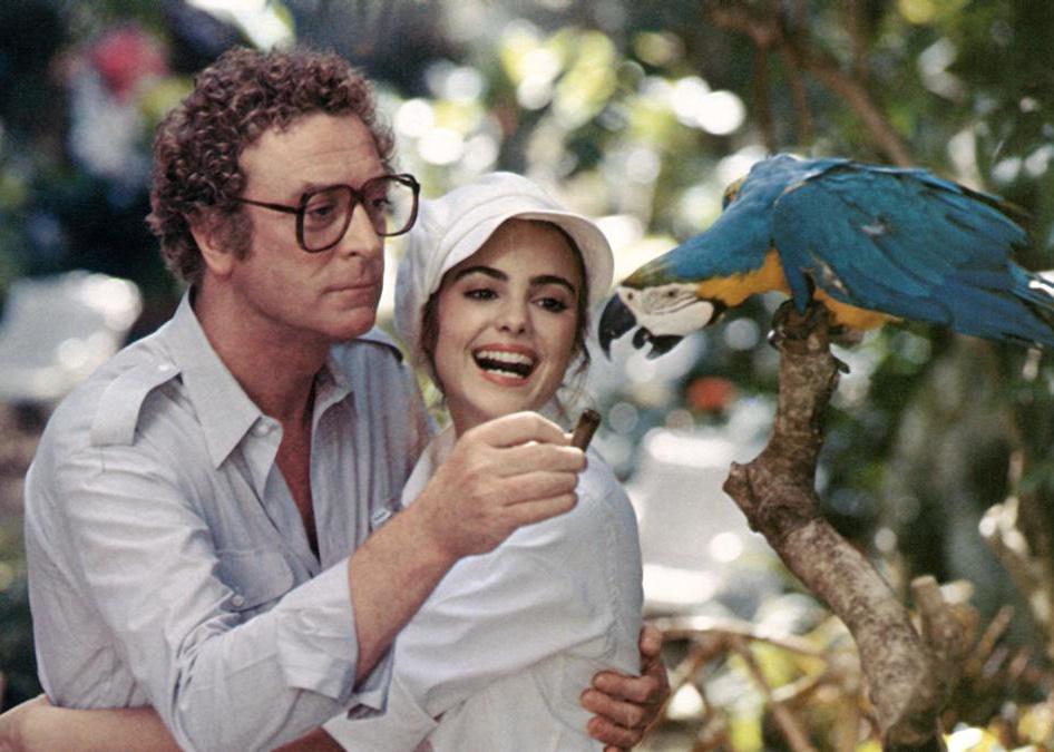 Michael Caine and a young woman laugh while feeding a parrot.
