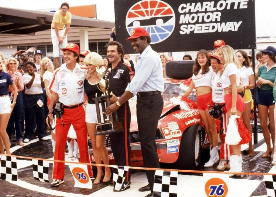 Burt Reynolds and Loni Anderson posing with race fans and pit crew in front of a racecar.