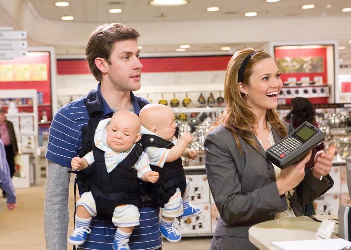 Mandy Moore holding a credit card machine in a store next to John Krasinski wearing a baby carrier holding two baby dolls.