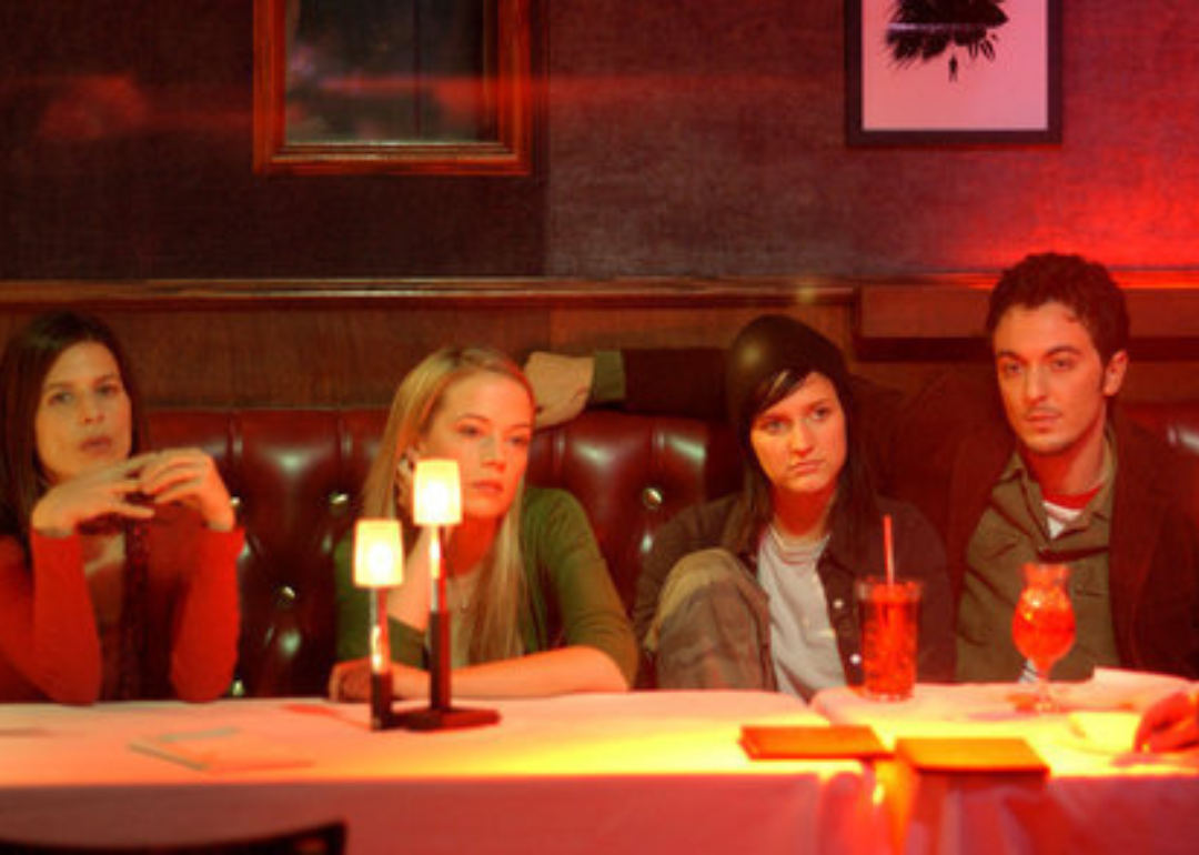 Three women and a man sit at a dimly lit table with blank stares.