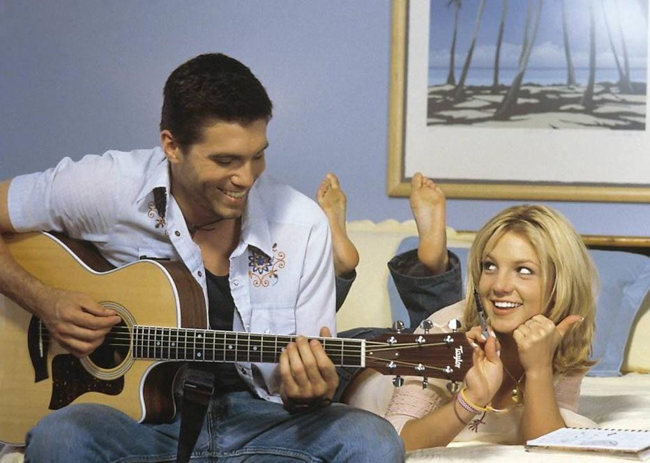 Britney Spears lays on a bed with a notebook next to a boy playing guitar.