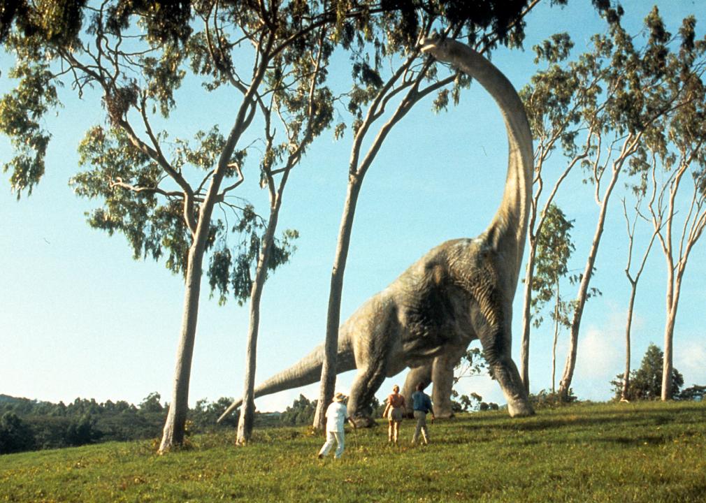Human characters from the movie gaze upward at a giant long neck dinosaur.
