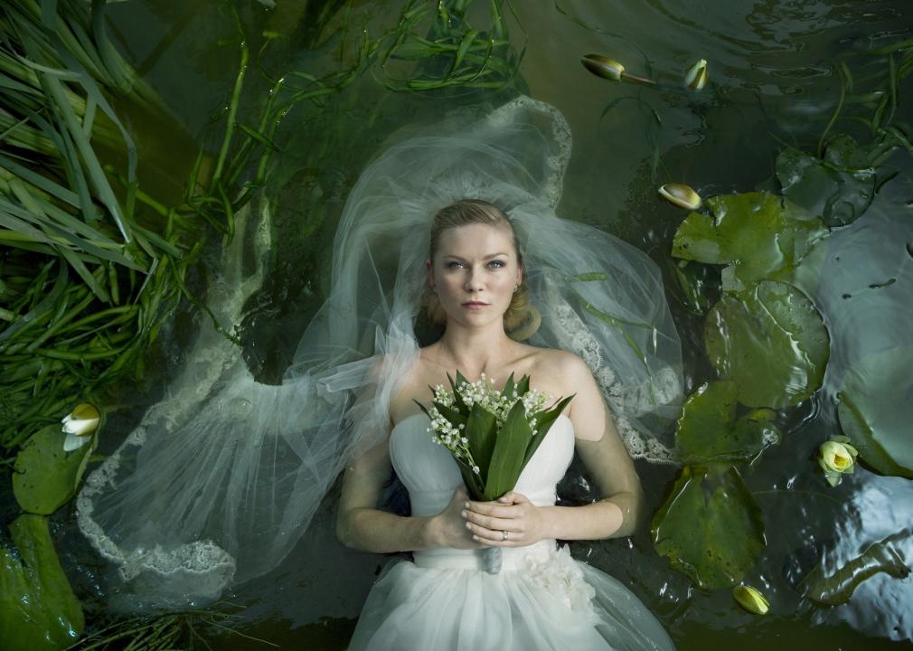 A woman in a strapless wedding gown and a blank stare floats in water among blossoming lily pads.