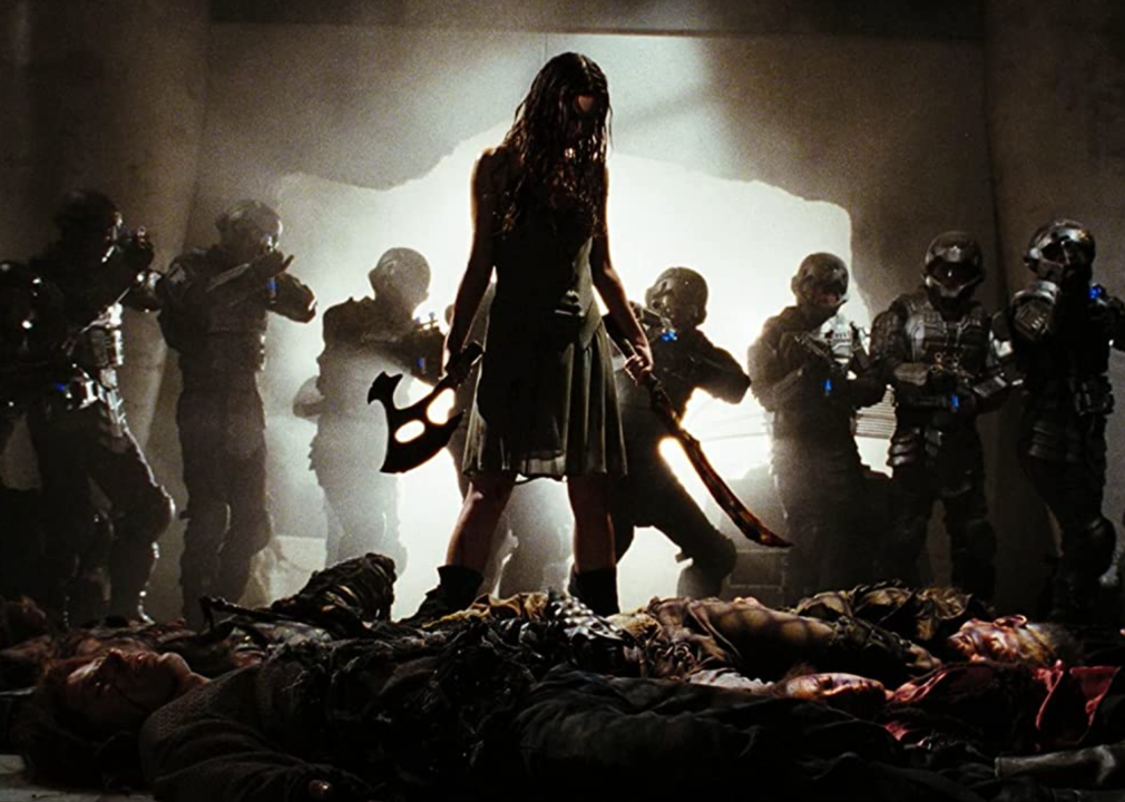 A woman with swords stands over the defeated while surrounded by robots aiming weapons at her.