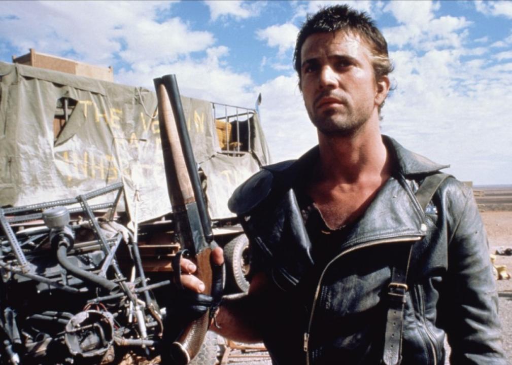 A rough looking man in a black leather jacket stands next to a torn up army truck and old vehicle parts.