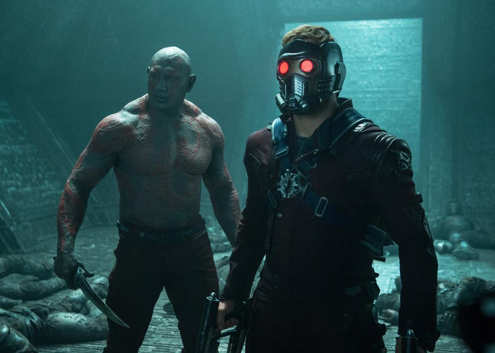 A muscular man with red drawings across his body and a knife stands next to a man with a face mask on with red lit eyes and a gun.