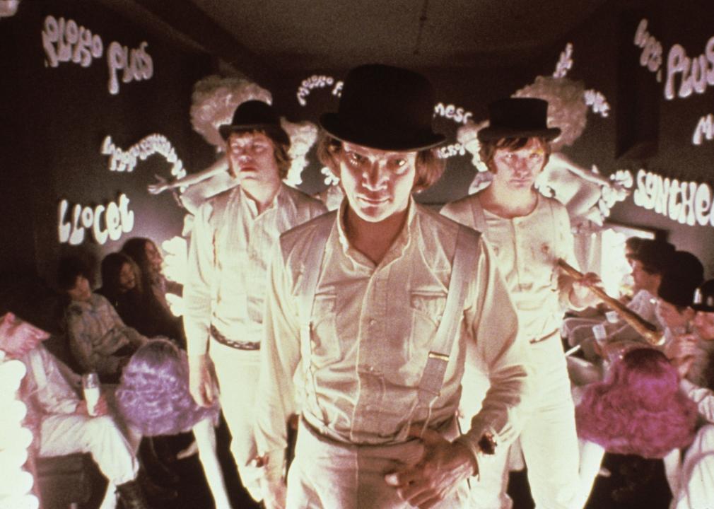 3 men dressed in all white with brown hats and baseball bats walk through a row of people sitting.