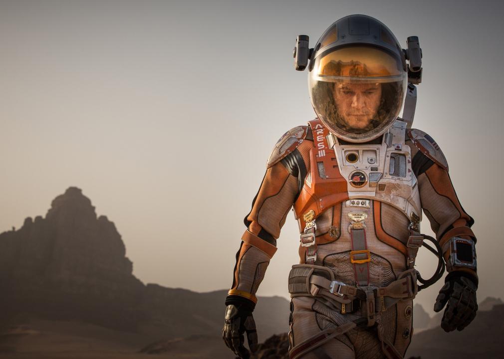 A man in an orange spacesuit walks on a desert-like planet.