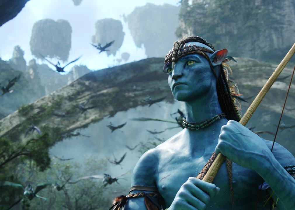 A blue being stands in a jungle with a pointy weapon.