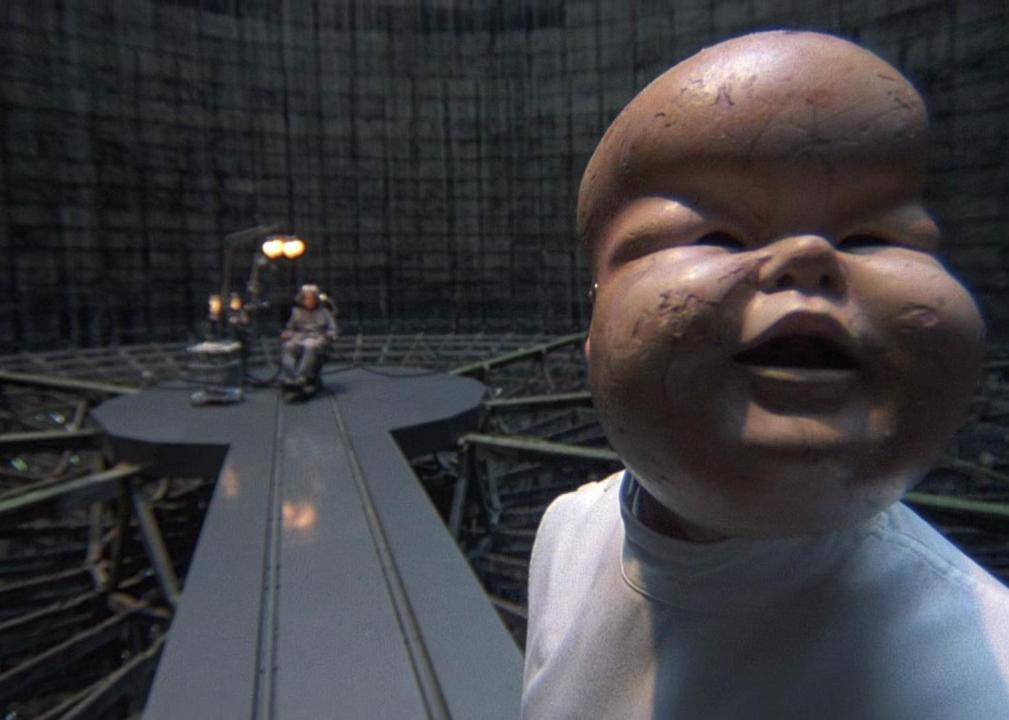Someone wearing a chubby baby mask and a seated man in background.