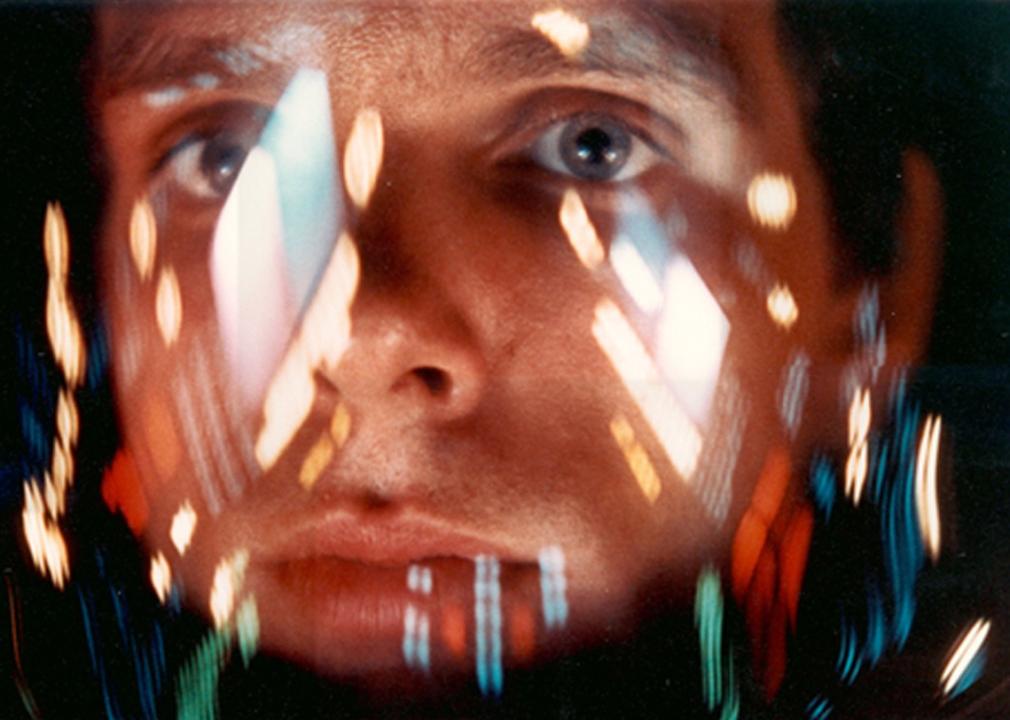 Man looking out of a space helmet with the colors from a machine's blinking lights reflecting all over his face shield.