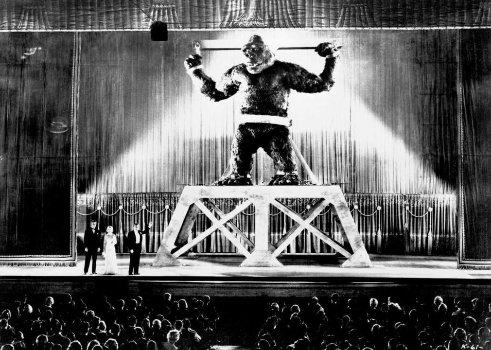 An enormous gorilla tied up on display on stage in front of a crowd with dressed up people standing next to him.