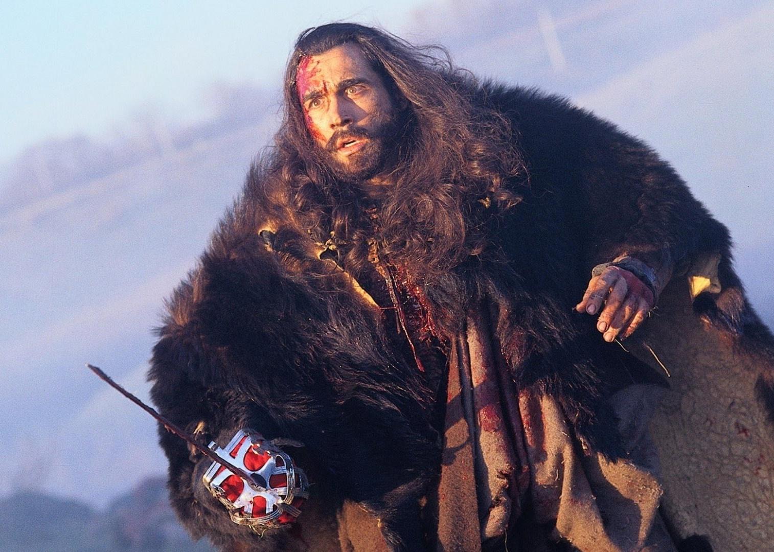 A man with long dark hair wearing a thick fur jacket and blood on his face holds a sword.