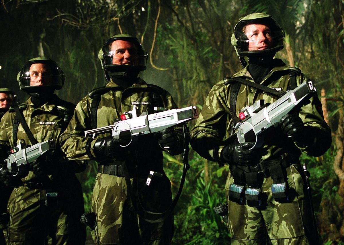 Edward Burns leads a group of men in camo suits, space helmets and guns.