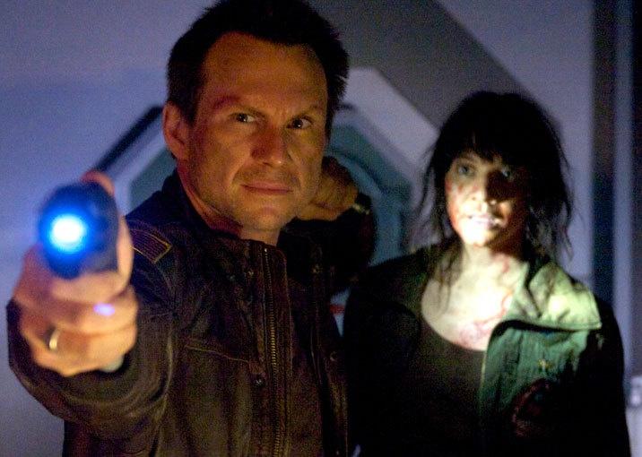 Christian Slater shines a blue light on something ahead while a woman stands behind him.