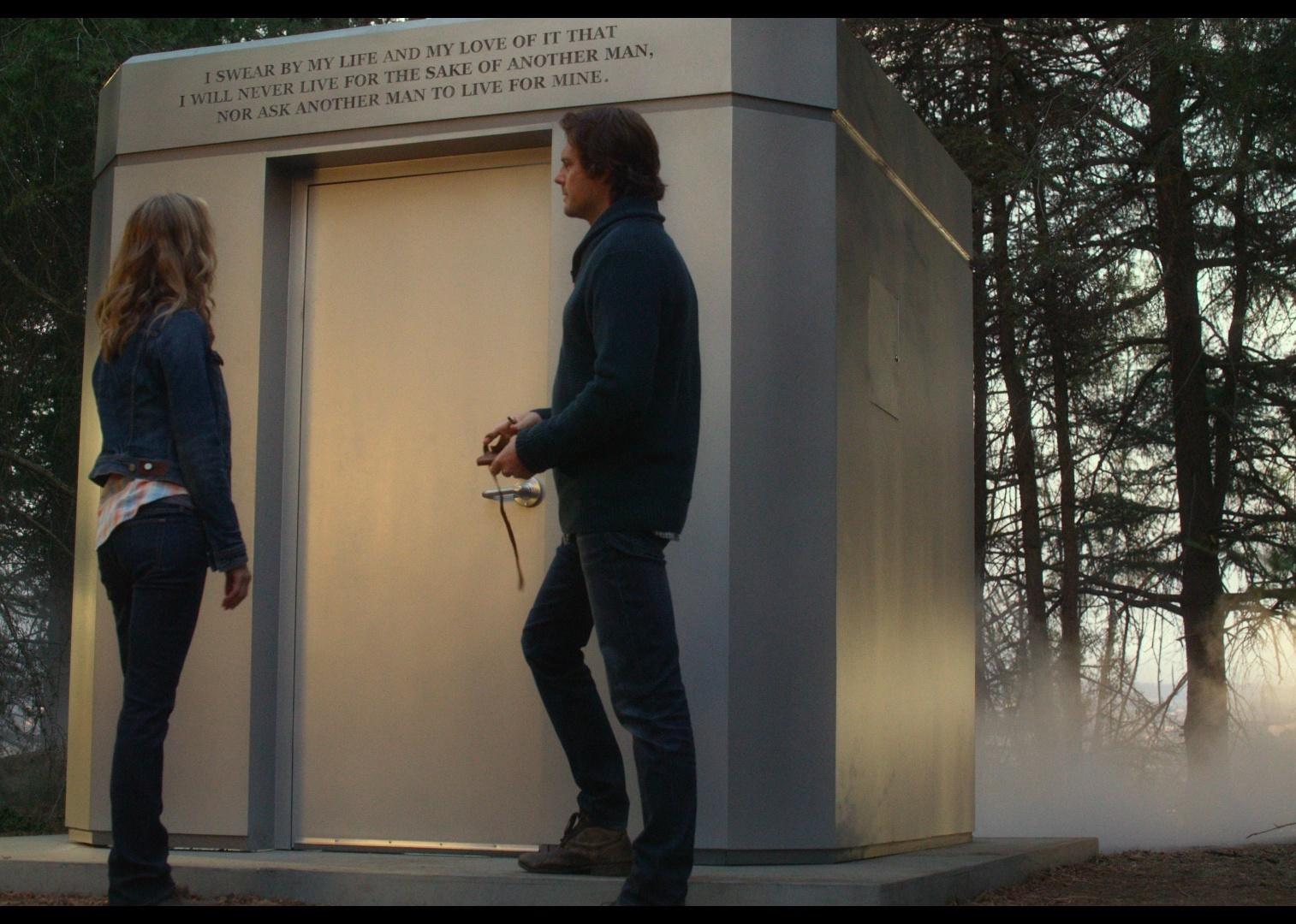 A man and woman stand in front of a giant square metal box in the woods with writing on the front.