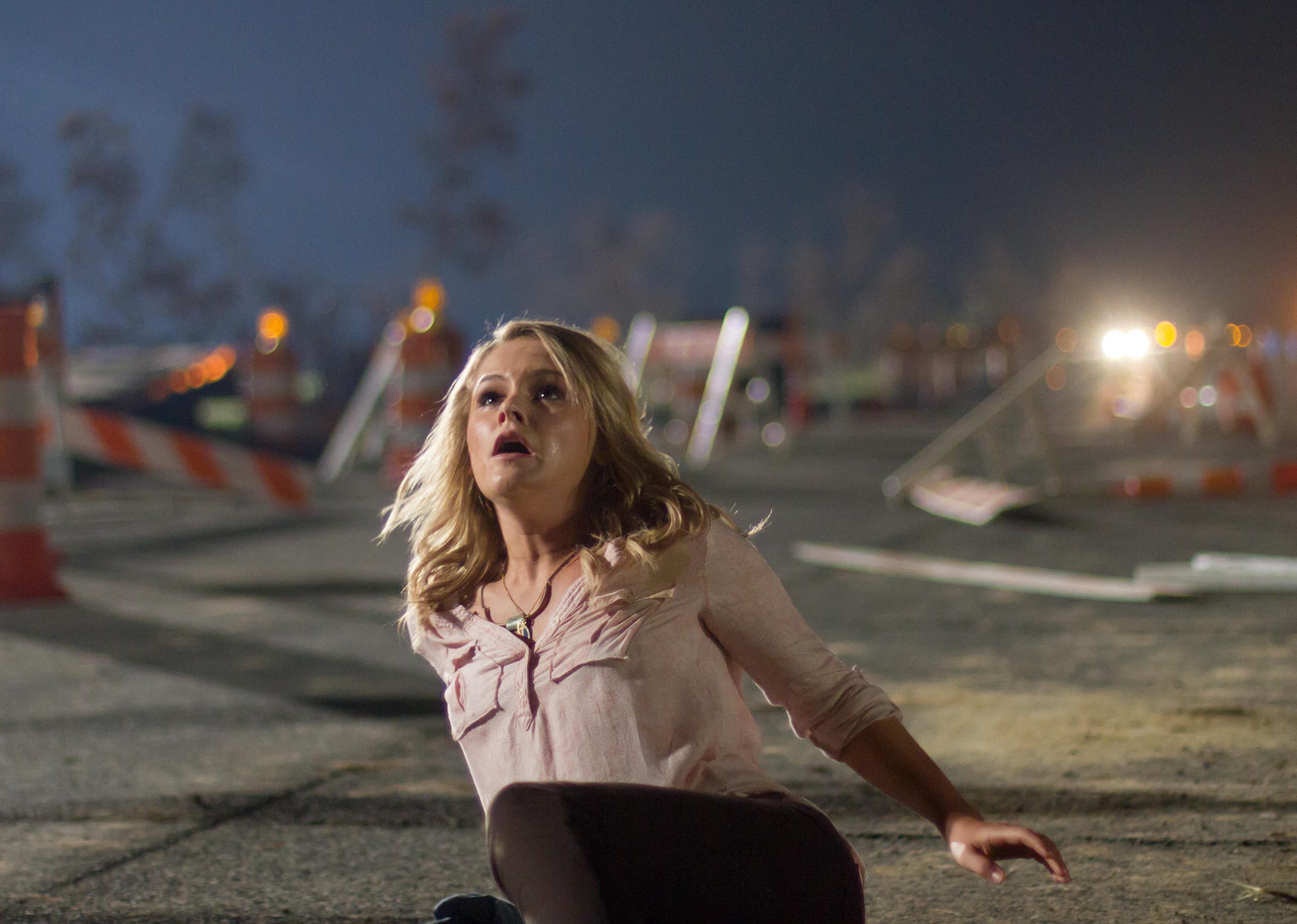 A blonde woman on the ground in chaotic wreckage looking up to the sky.