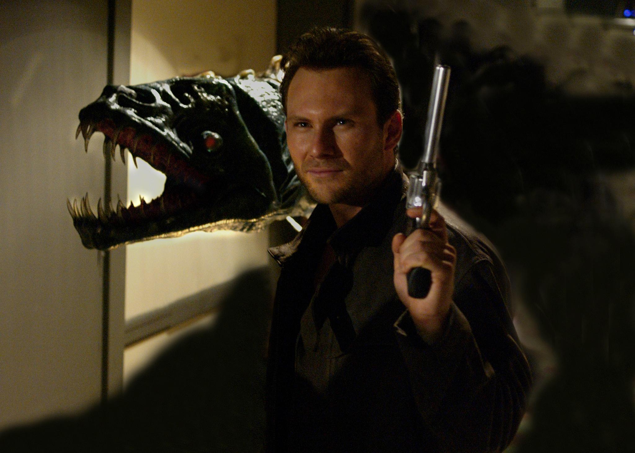 Christian Slater holding a pistol and standing in front of a dinosaur.