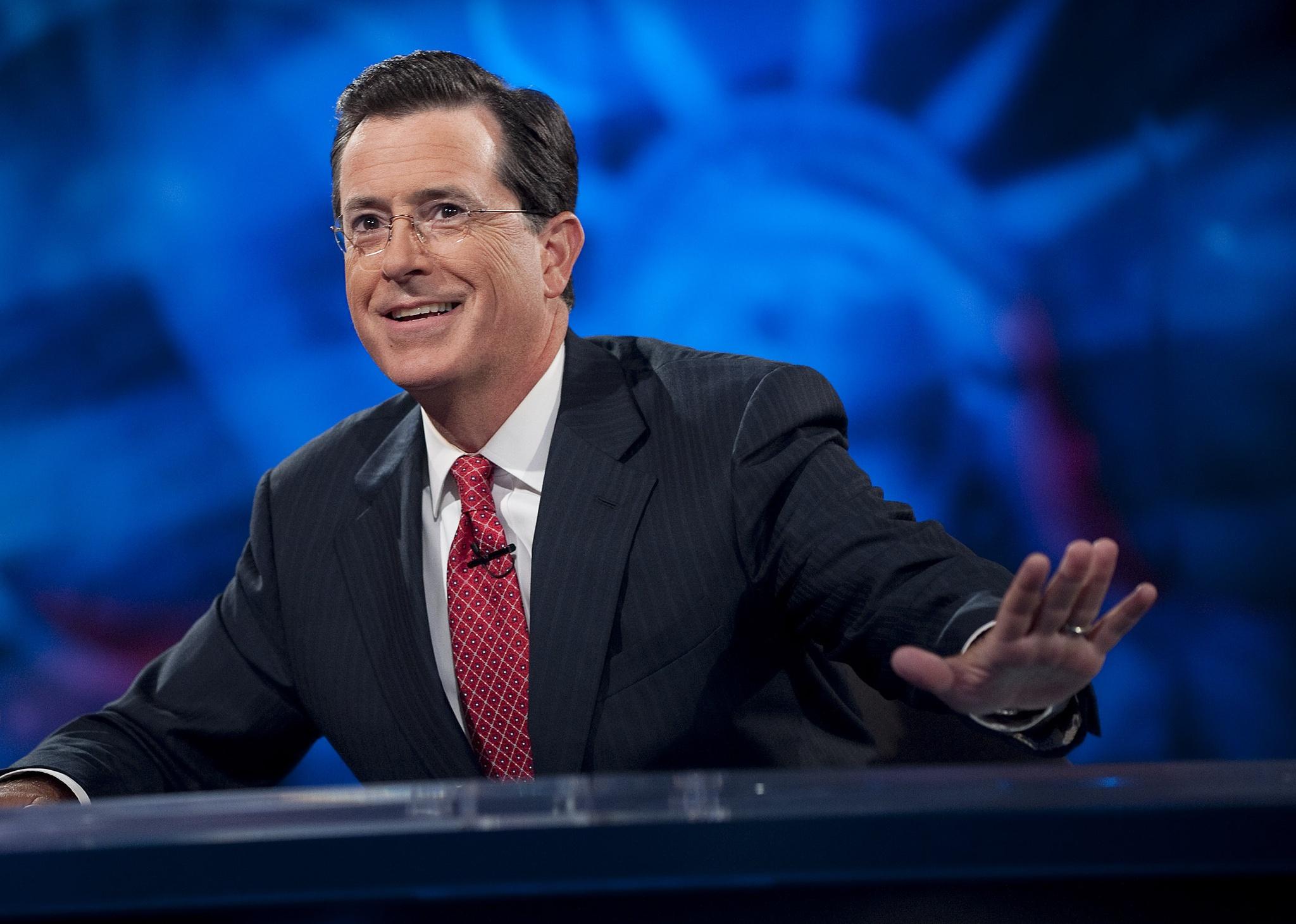 Stephen Colbert smiling at a desk in front of a blue background.