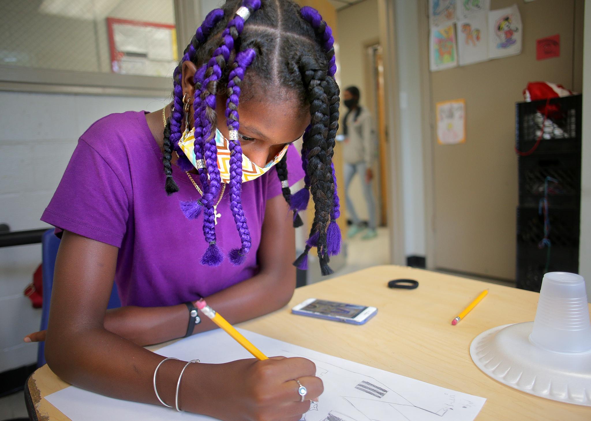 A girl wearing a mask draws with a pencil at a desk.