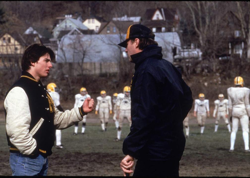A guy in a letter jacket talking to a coach while a suited up football team waits in the background.