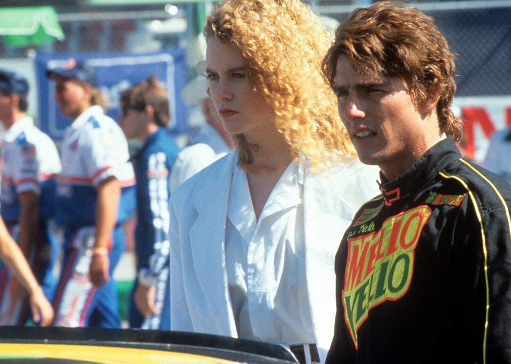 Tom Cruise, dressed in a race car driver uniform, stands next to Nicole Kidman.