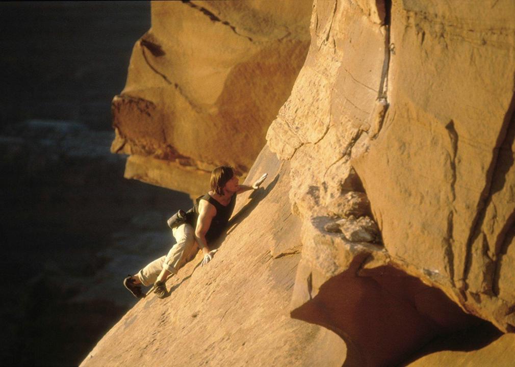 A man with shoulder length black hair climbs up the face of a rock at the top of a mountain.