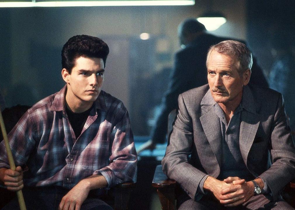 Tom Cruise and Paul Newman sit with serious looks on their faces next to a pool table.