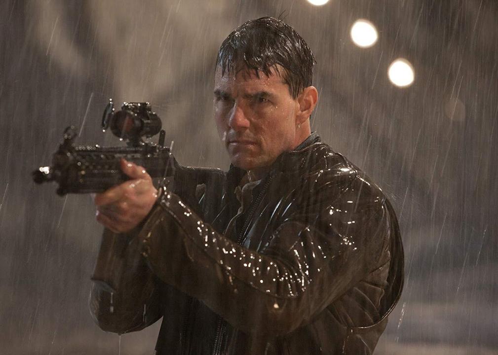 Tom Cruise stands in the pouring rain while pointing a large gun.