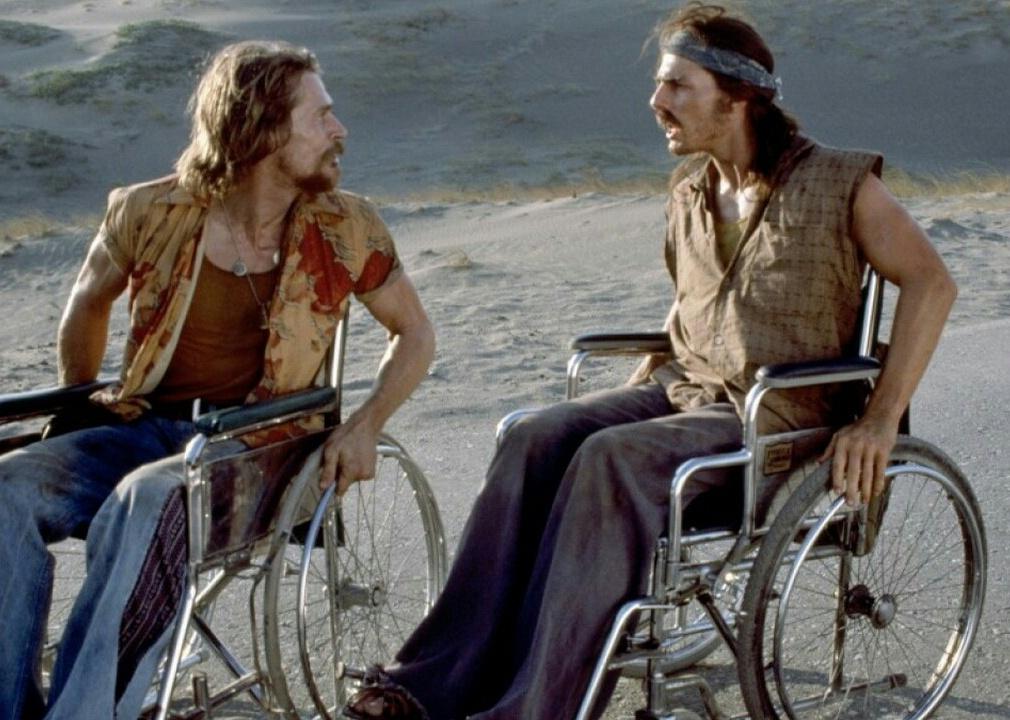 Two men in wheelchairs wearing tattered clothing argue on the beach.