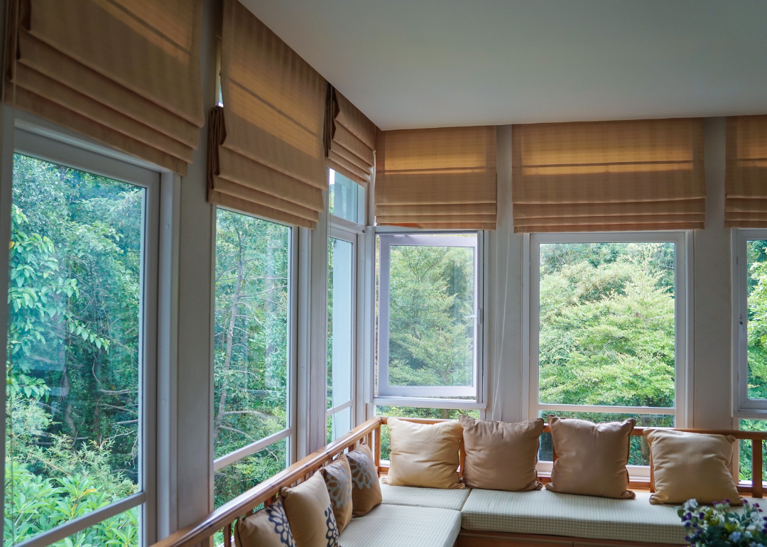 Living room windows facing the forest covered with brown roman shades.
