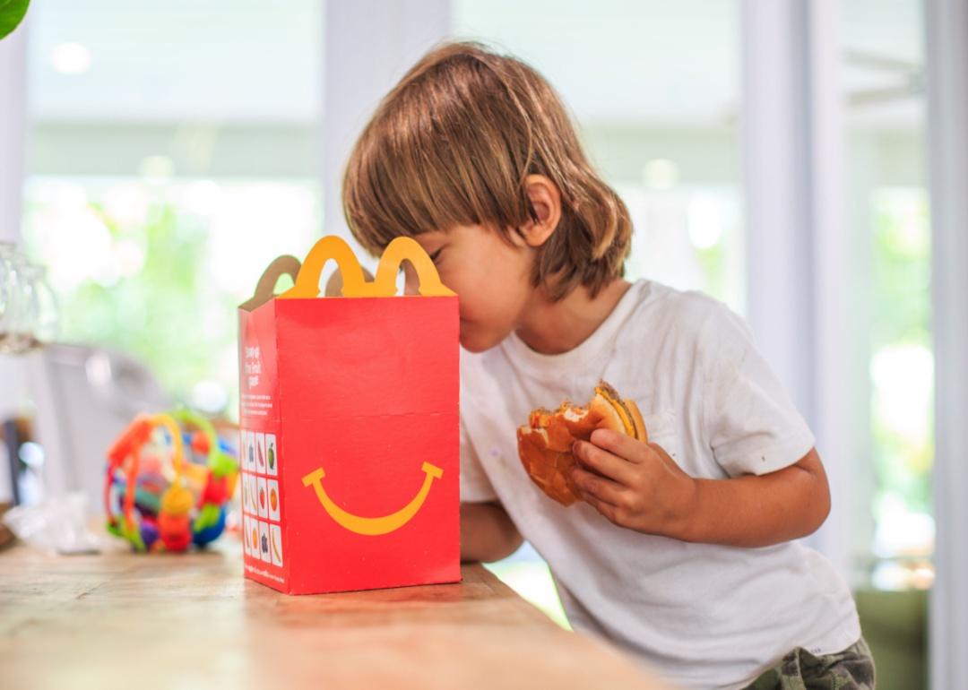 A young boy looking inside a red McDonald's happy meal box while eating a cheeseburger.