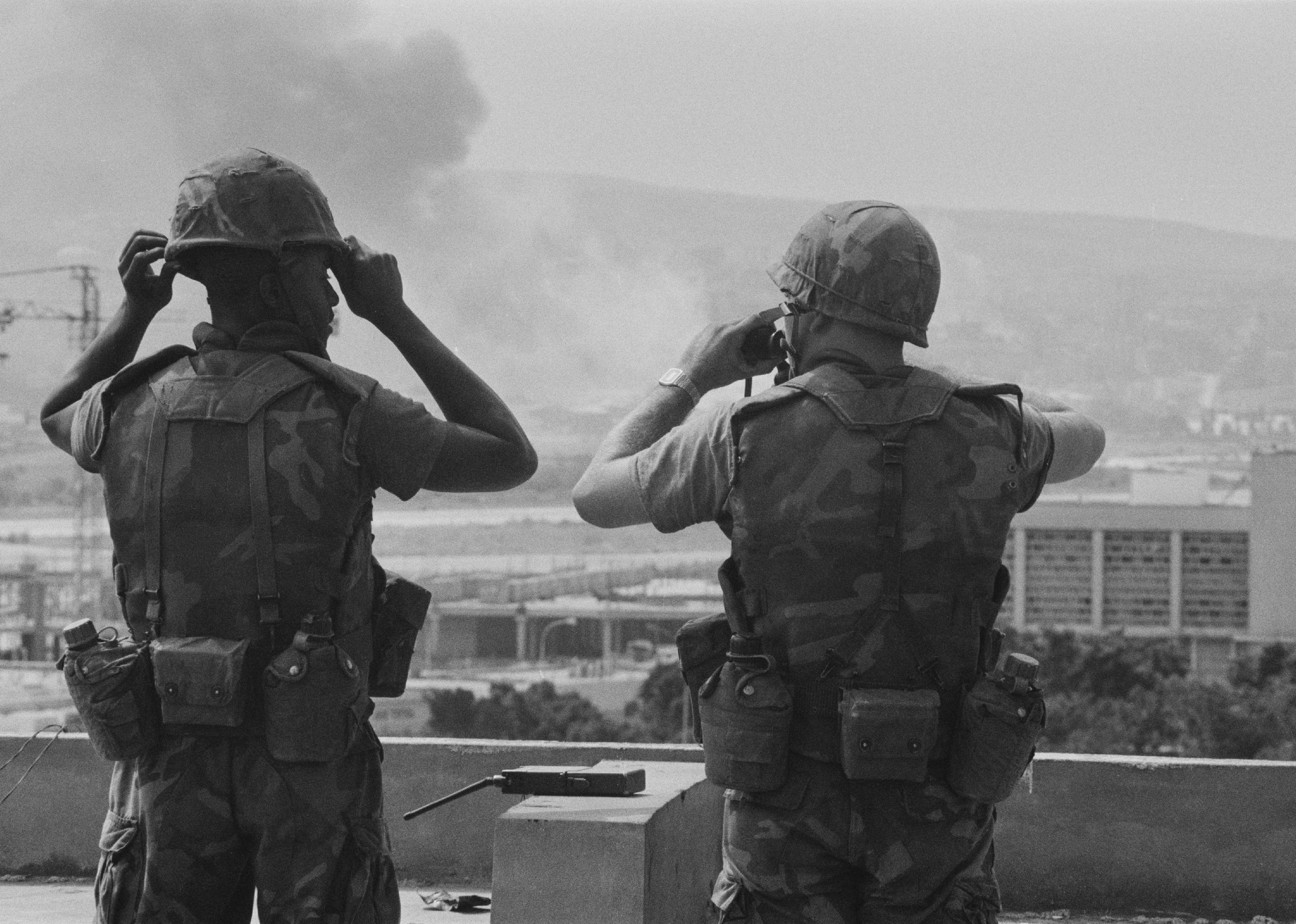 Marines stand guard on top of a building with smoke in the distance.