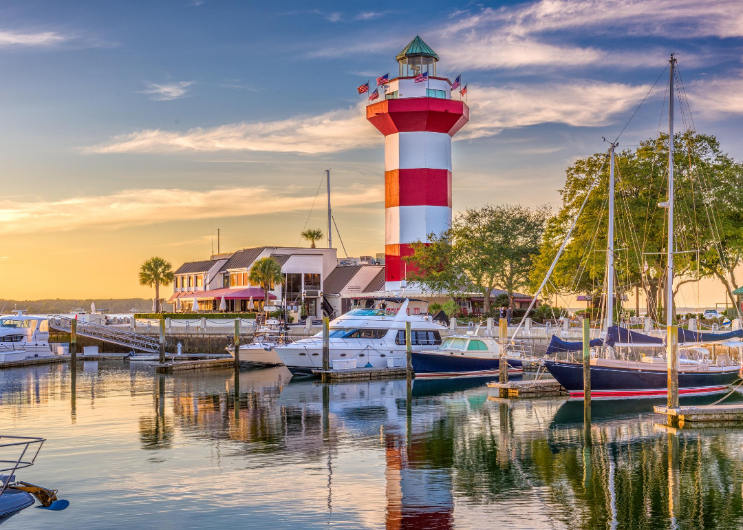 Red and white striped lighthouse and boats in marina at sunrise.