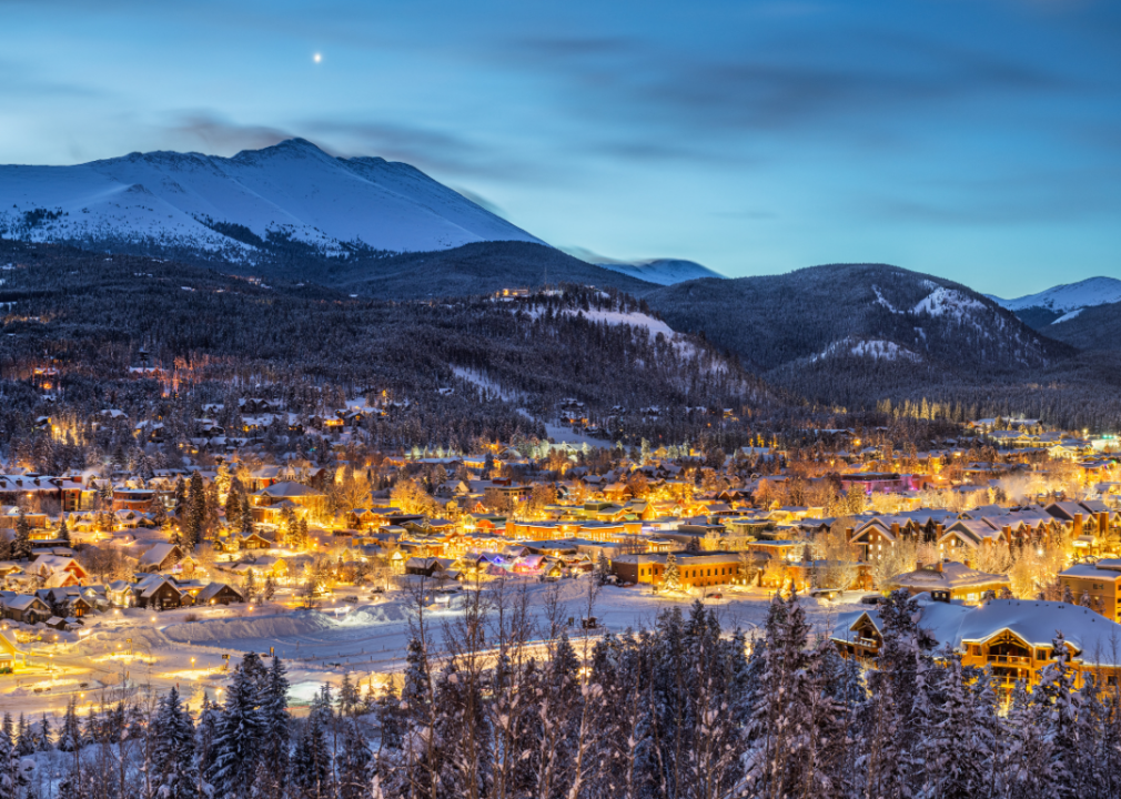 A snow-covered, warmly lit town with large mountains in the background in the evening. 