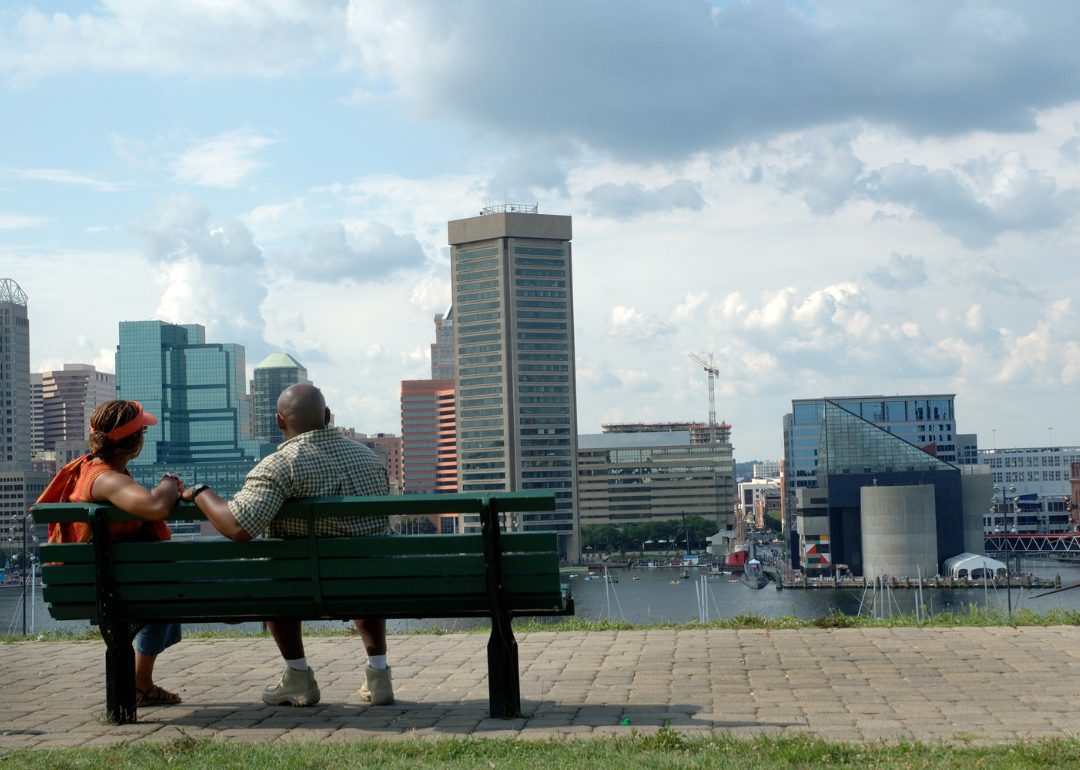 A couple sitting on a park bench overlooking the water and the Baltimore skyline.