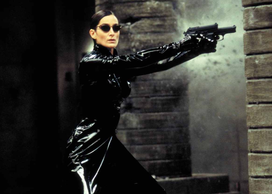 Carrie-Anne Moss in all black pointing a gun.