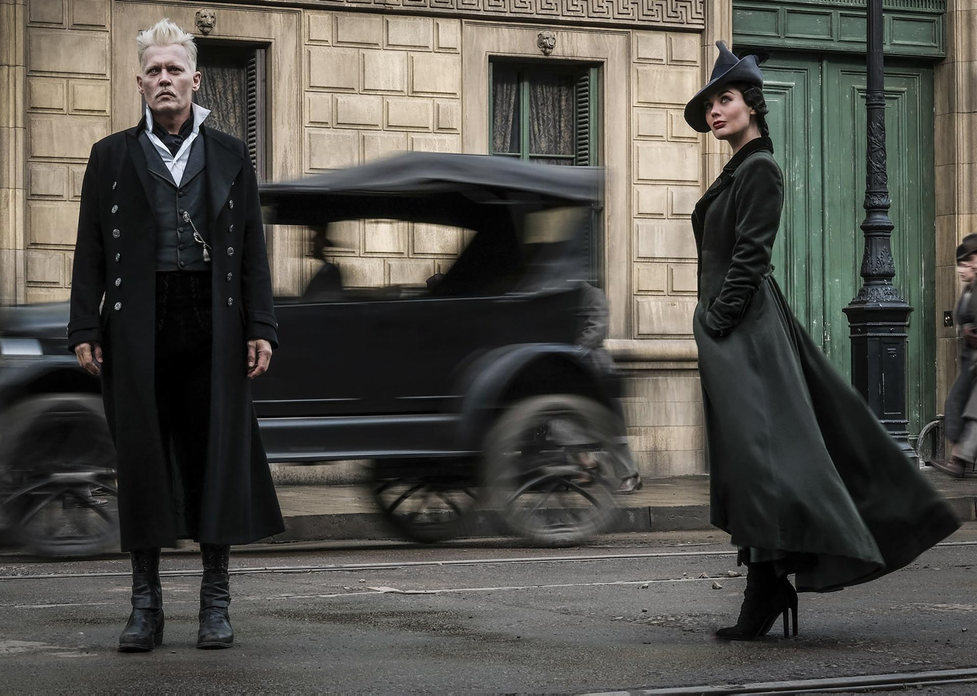 Johnny Depp with white hair and Poppy Corby-Tuech in all black standing on a street with an old car whizzing by behind them.