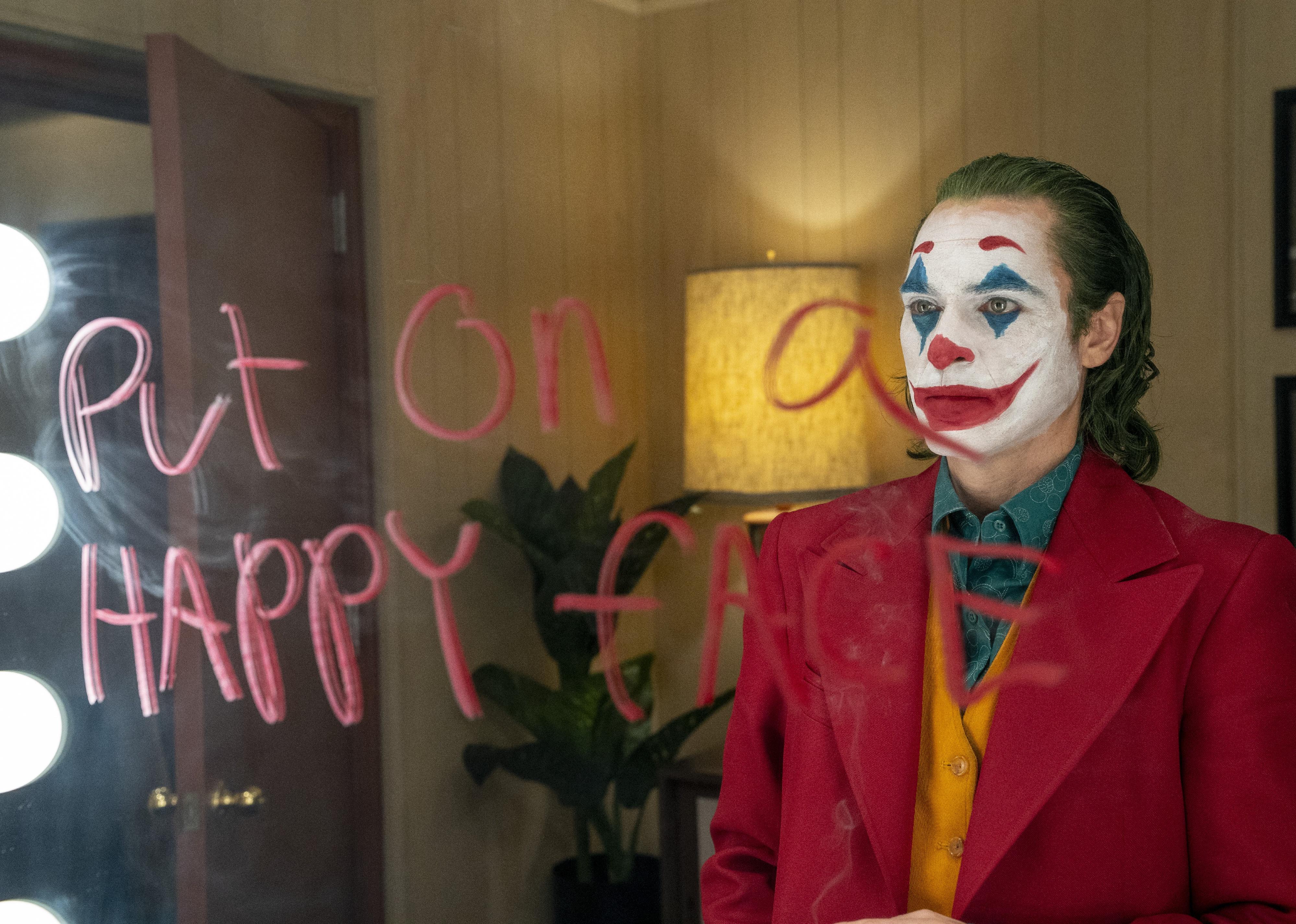 Joaquin Phoenix with clown makeup on in a red jacket looking in a mirror.