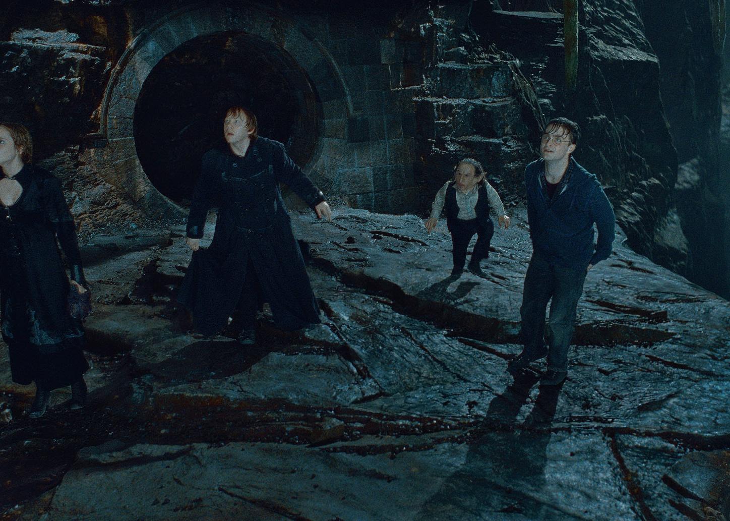 Warwick Davis, Rupert Grint, Daniel Radcliffe, and Emma Watson standing outside of a tunnel on a cliff.