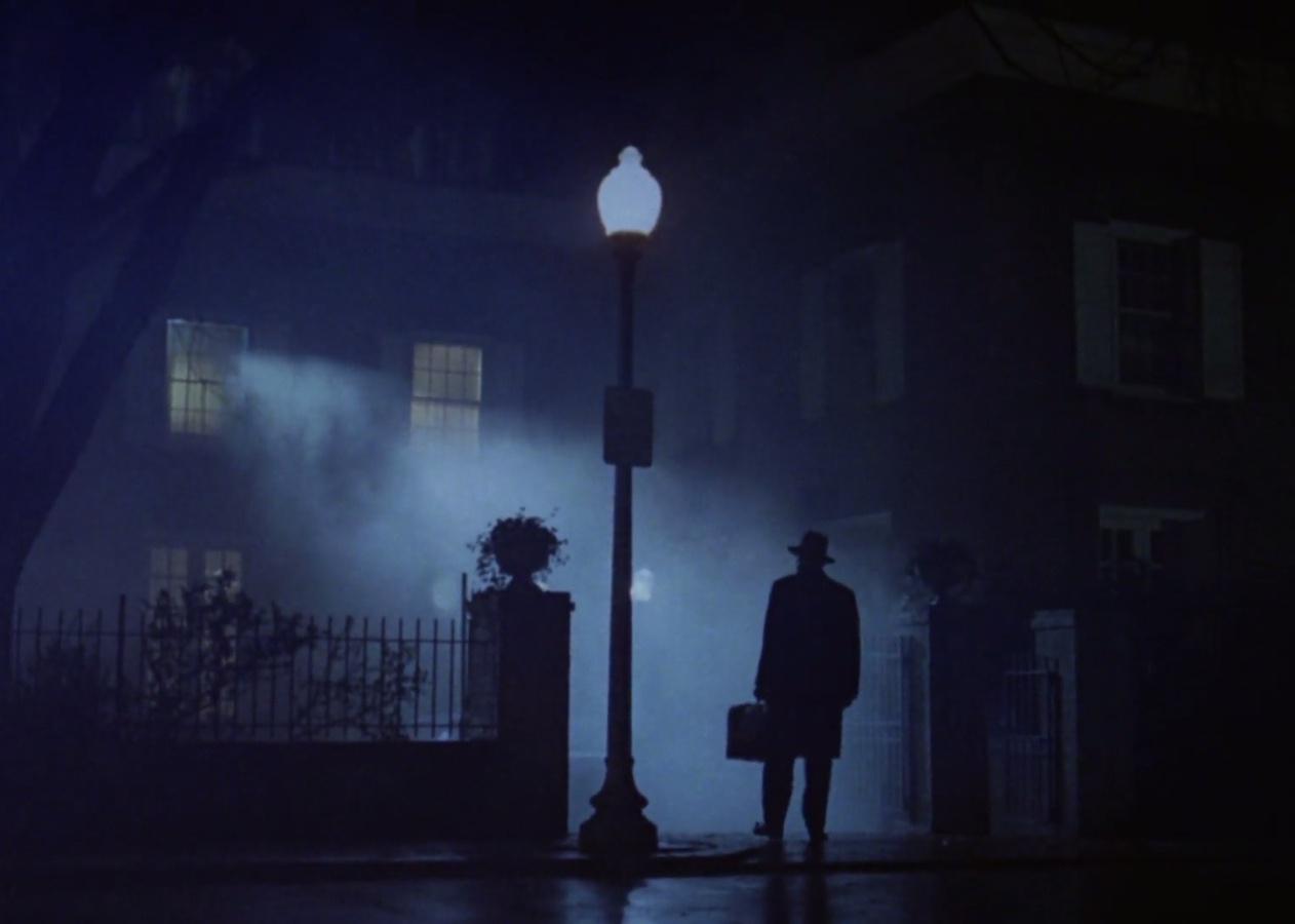 A silhouette of a man in a hat standing in front of a house on a foggy night.