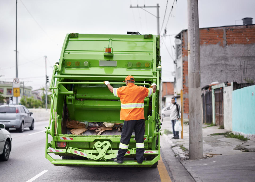 A trash collector riding on the back of a green garbage truck.
