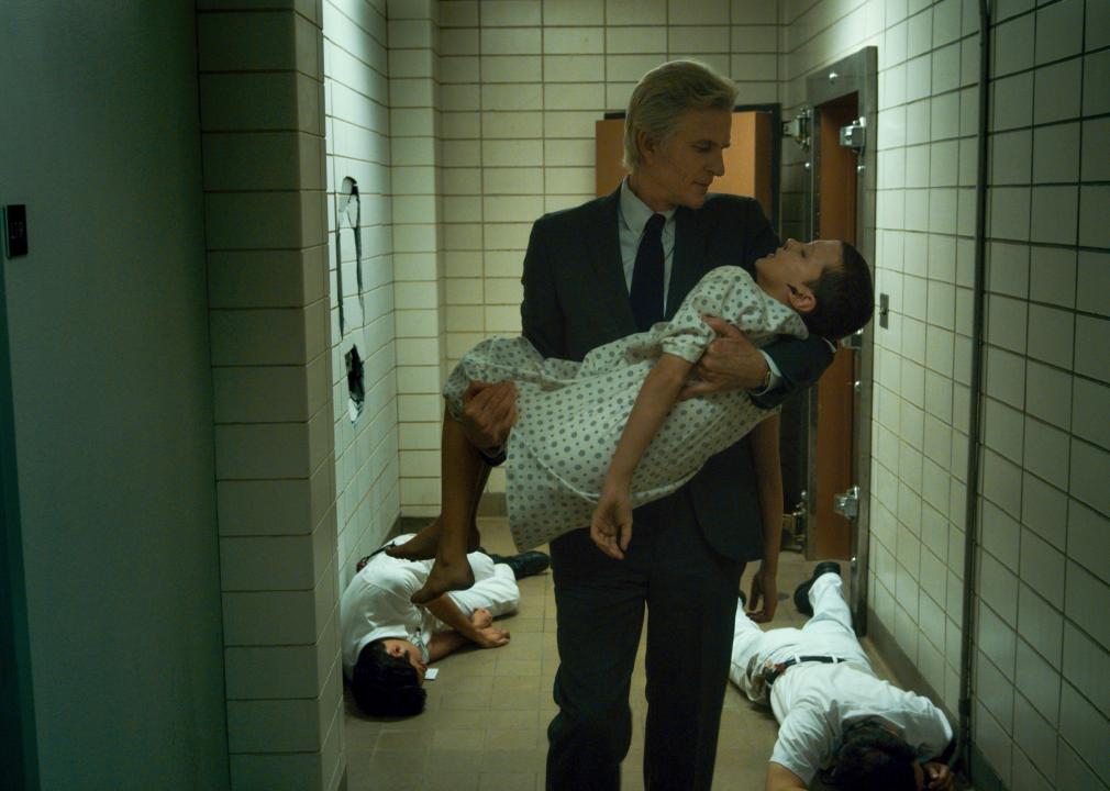 Matthew Modine carrying Millie Bobby Brown in a hospital gown past collapsed people on the floor.