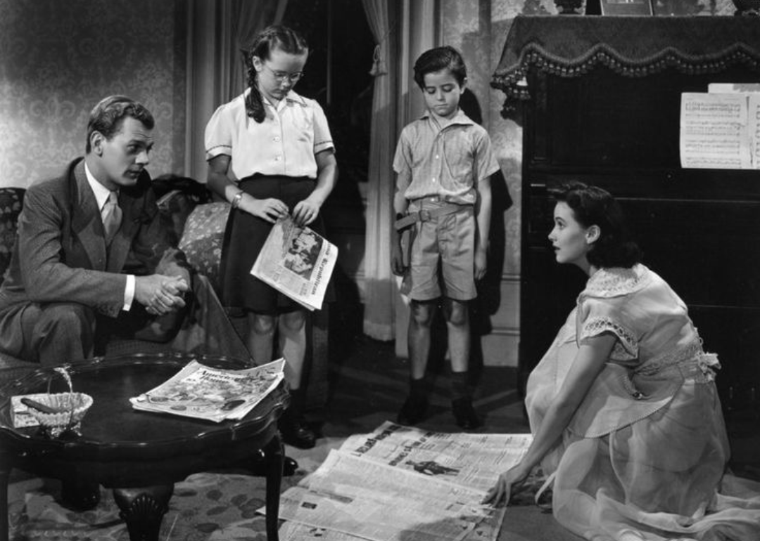 A family sits in the living room together looking at a newspaper scattered on the floor.